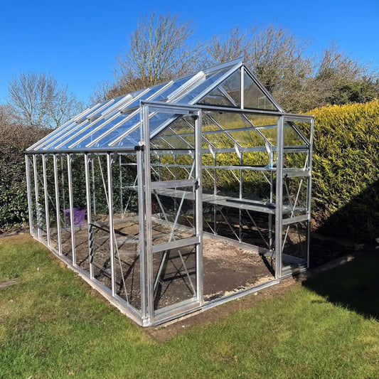 A glass Rhino Greenhouse with an aluminum frame stands empty on a grassy lawn. The surrounding environment features green hedges and bare trees under a clear blue sky.