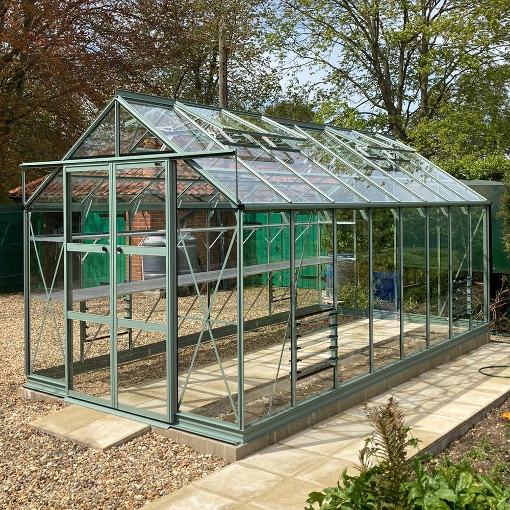 A glass Rhino Greenhouse stands on a tiled base, surrounded by gravel and garden foliage, with large windows reflecting sunlight, and trees visible in the background.