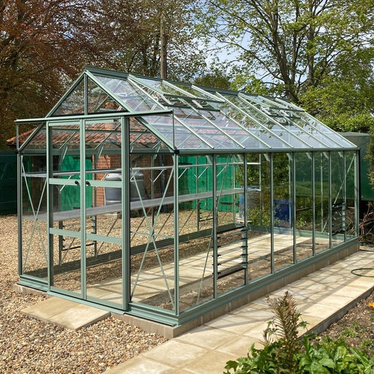 A glass Rhino Greenhouse stands on a tiled base, surrounded by gravel and garden foliage, with large windows reflecting sunlight, and trees visible in the background.