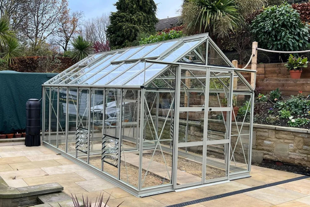 A glass Rhino Greenhouse with a metal frame stands on a patio. It's surrounded by a garden with various plants and a stone retaining wall.