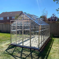 A glass Rhino Greenhouse stands on a sunny lawn, surrounded by a wooden fence and nearby house, reflecting bright sunlight on its transparent panels.