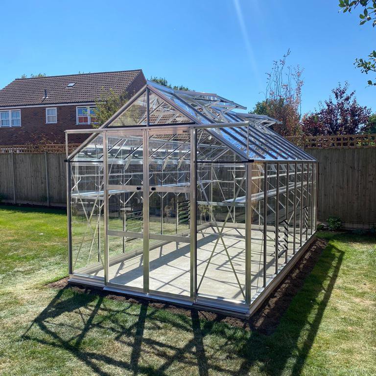 A glass Rhino Greenhouse stands on a sunny lawn, surrounded by a wooden fence and nearby house, reflecting bright sunlight on its transparent panels.