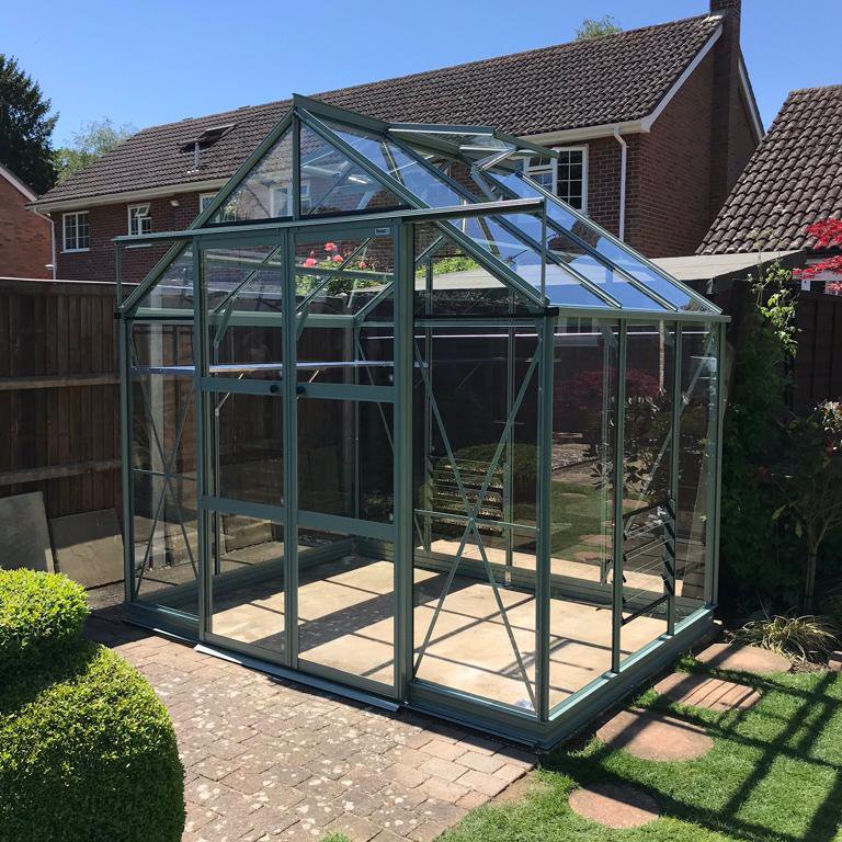 A glass Rhino Greenhouse stands on a brick patio, with metal frames, surrounded by a wooden fence and a well-maintained garden in a residential backyard setting.