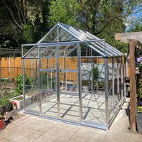 A glass and metal-framed Rhino Greenhouse stands on a patio, surrounded by garden plants and a wooden fence, under a clear blue sky with trees in the background.