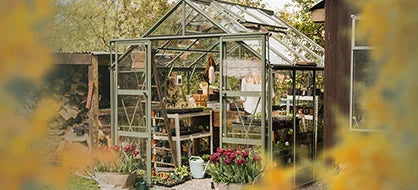 A glass Rhino Greenhouse shelters gardening tools and plants, standing amidst a garden setting with flowers and a nearby building. The environment is lush and verdant.