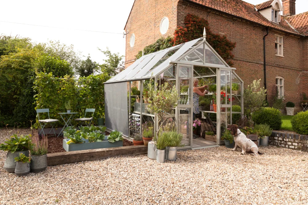 A glass Rhino Greenhouse contains a person tending plants inside. Gravel paths and potted plants surround it, with a dog sitting nearby. A brick house and garden are in the background.