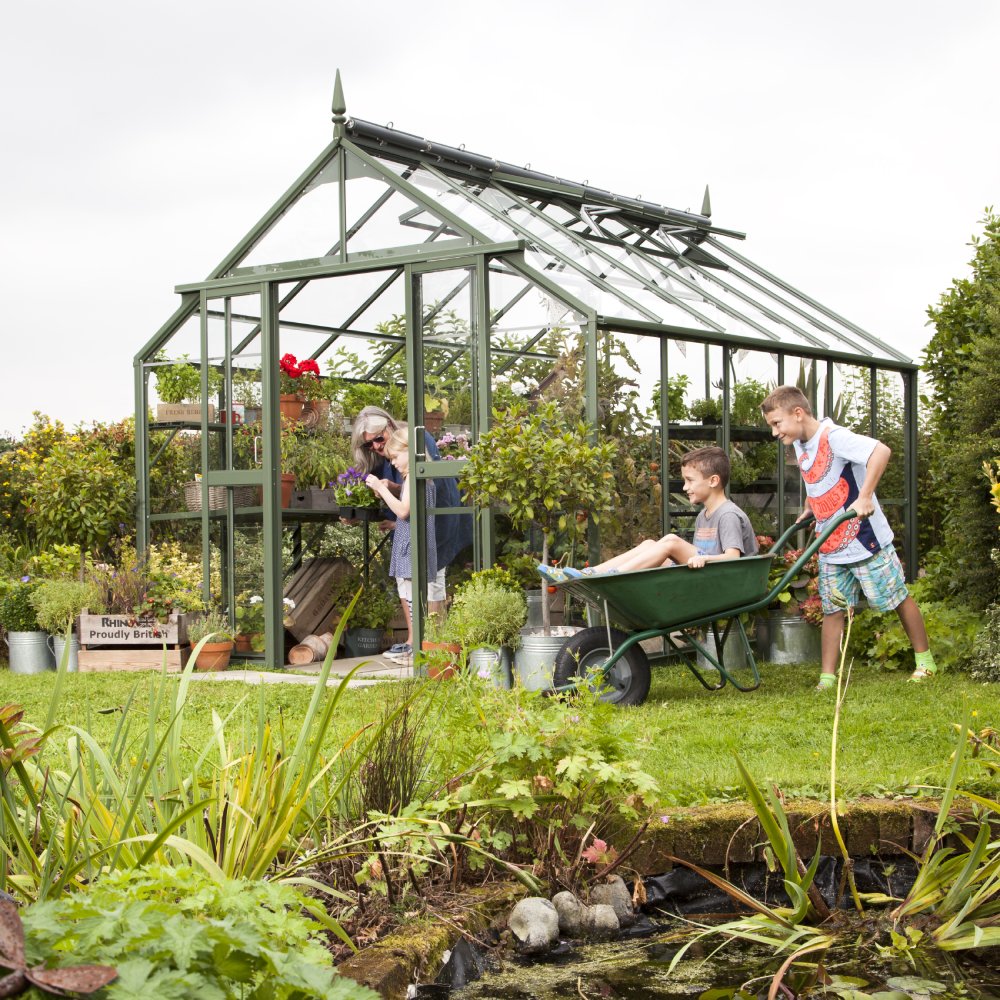 A green Rhino Greenhouse filled with plants and gardening tools stands in a lush garden. A woman tends to plants while two boys play with a wheelbarrow. Text: Rhino – Proudly British.