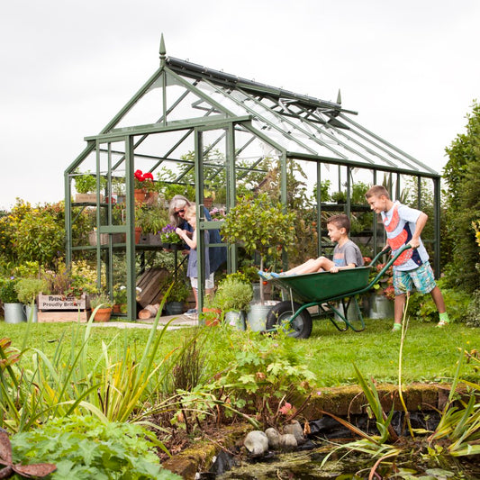 A green Rhino Greenhouse filled with plants and gardening tools stands in a lush garden. A woman tends to plants while two boys play with a wheelbarrow. Text: Rhino – Proudly British.