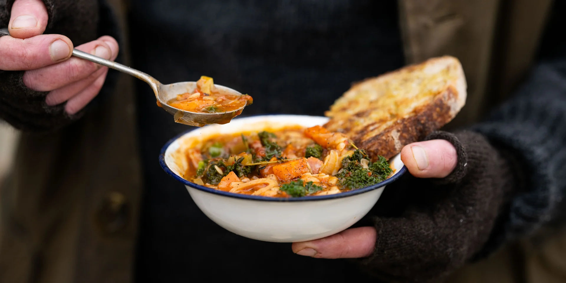 A person holds a bowl of vegetable stew with a slice of bread; another hand lifts a spoonful, set against a dark, blurred background.