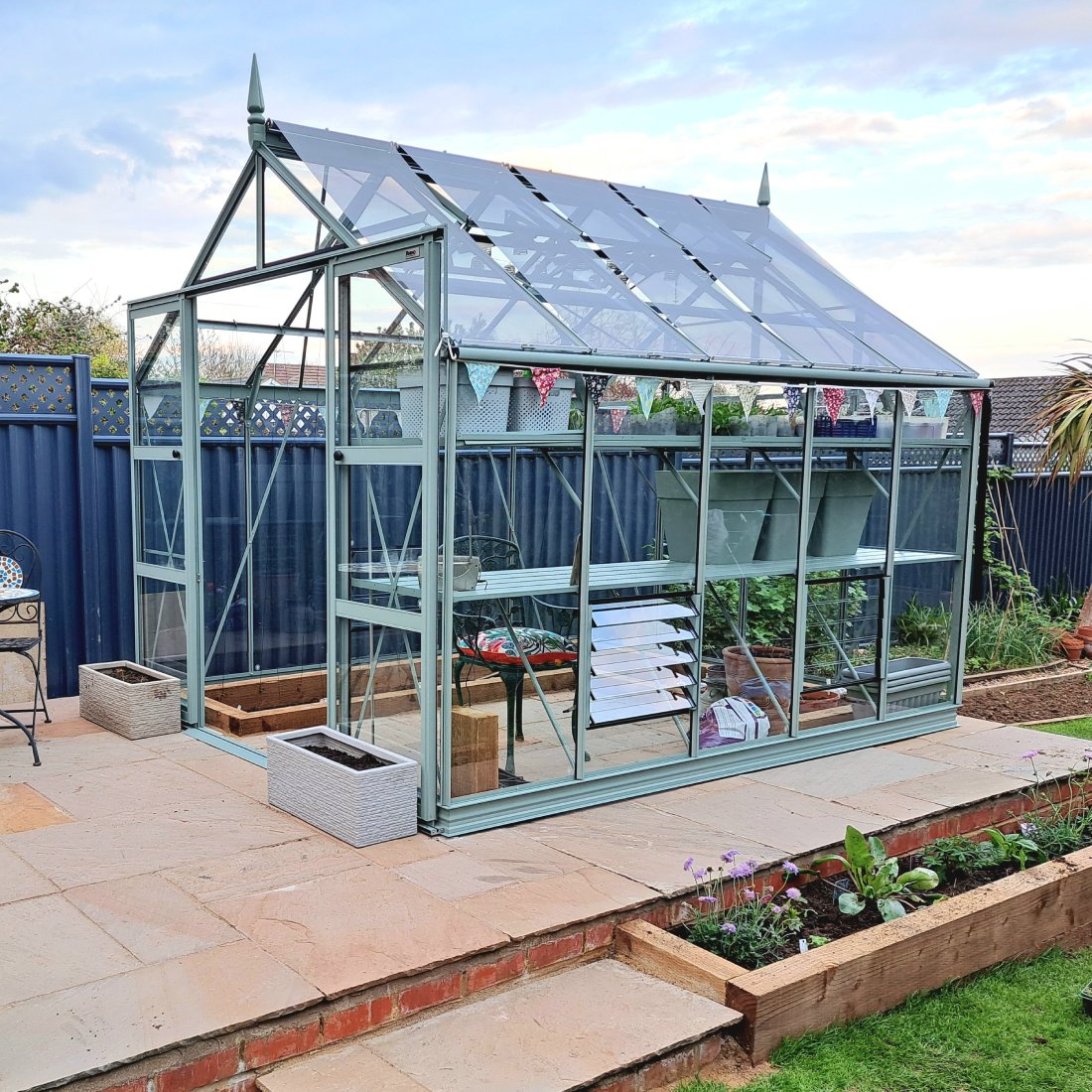 Rhino Greenhouse stands on a raised patio, featuring glass panels and metal frame. Inside, shelves hold gardening supplies. Outdoors, bordered by a blue fence, garden beds, and a cloudy sky.