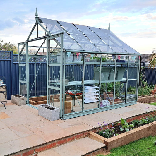 Rhino Greenhouse stands on a raised patio, featuring glass panels and metal frame. Inside, shelves hold gardening supplies. Outdoors, bordered by a blue fence, garden beds, and a cloudy sky.