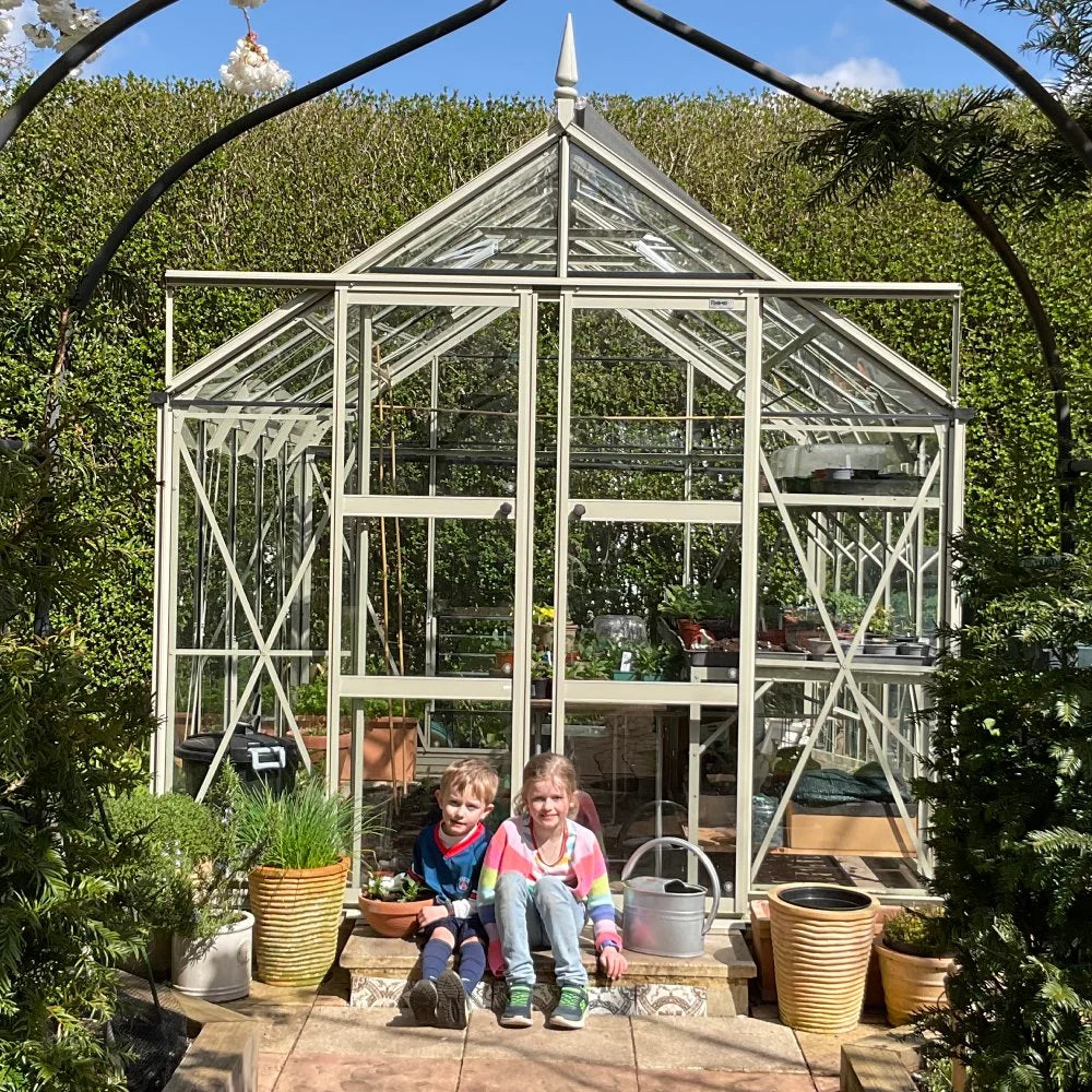 A glass Rhino Greenhouse stands amid lush greenery, housing various plants. Two children sit smiling in front, holding a bowl, surrounded by potted plants and a watering can.