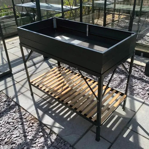 A rectangular, elevated garden bed stands on metal legs with a slatted wooden shelf beneath. It is placed on a tiled patio inside a glass Rhino Greenhouse.