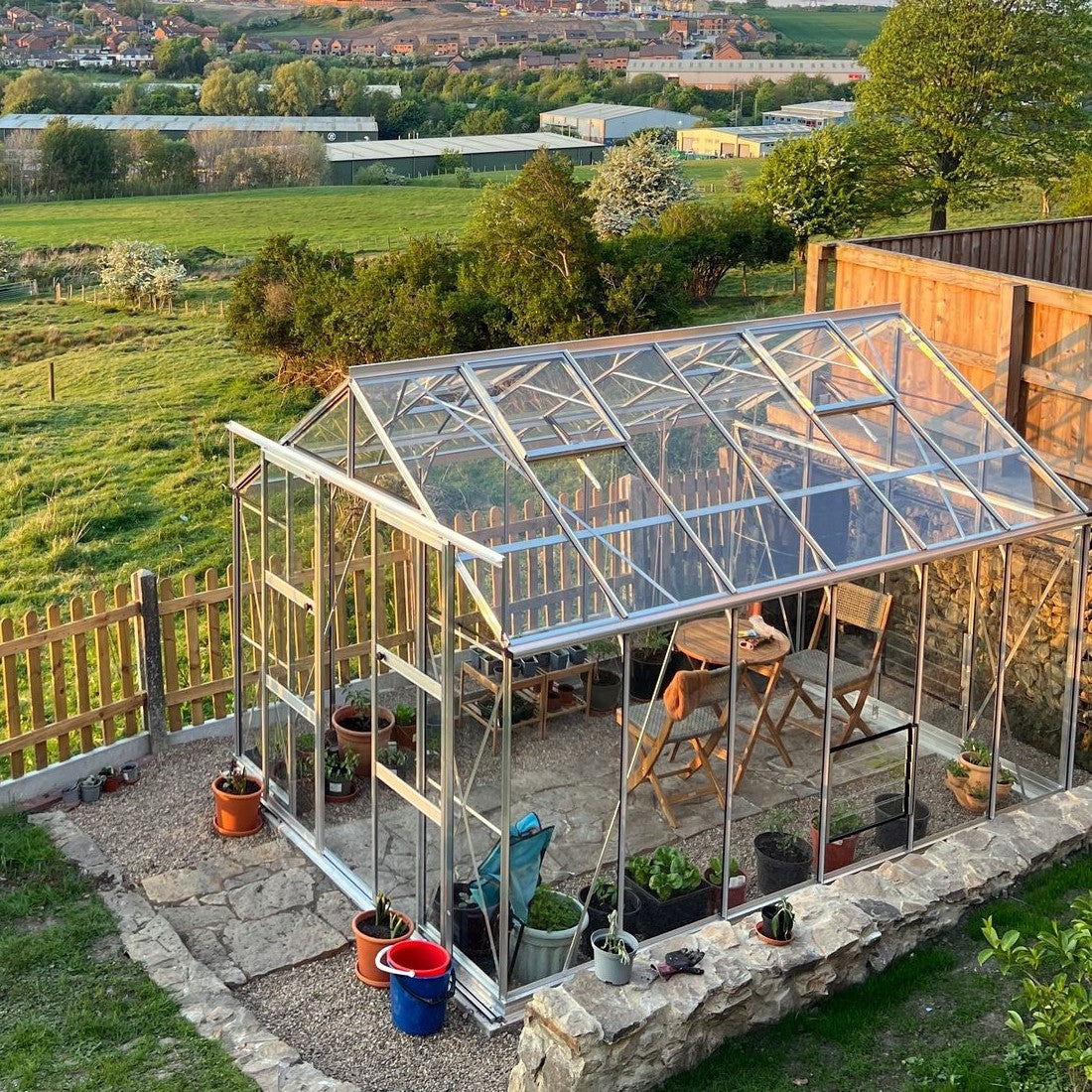A transparent glass Rhino Greenhouse houses potted plants and chairs, situated on a stone patio. It's surrounded by a wooden fence and overlooks a lush, expansive countryside landscape.
