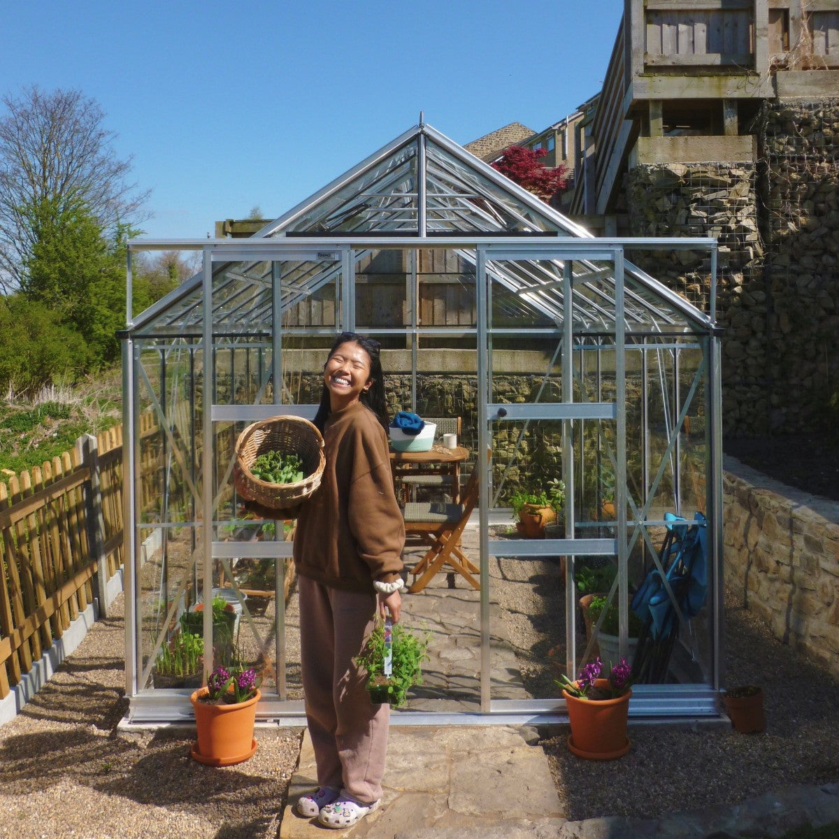 A person holds a basket of vegetables and plants, smiling in front of a glass Rhino Greenhouse surrounded by potted plants and wooden fencing, under a clear blue sky.