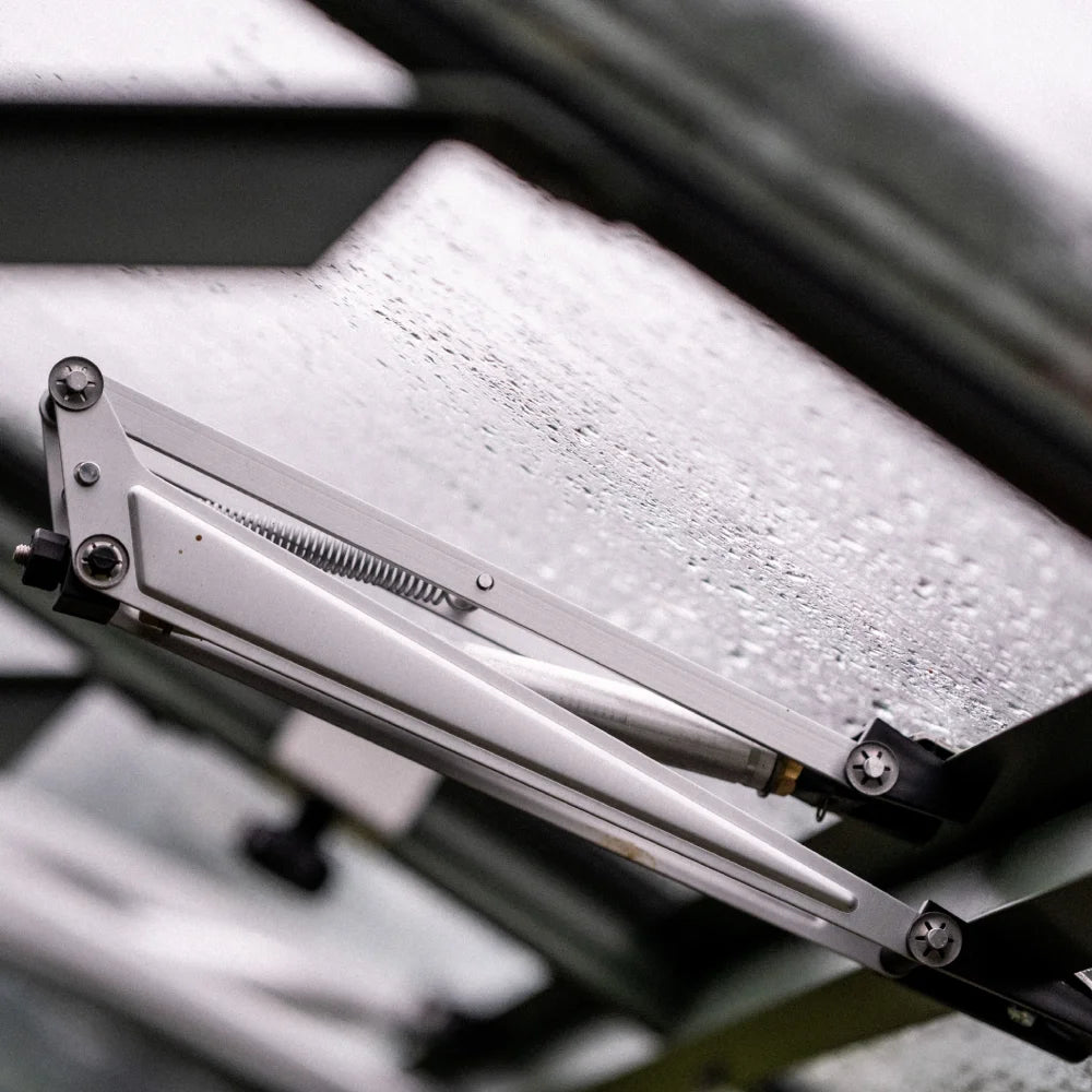 A hydraulic arm supports a rain-splattered glass pane, partially open, amidst the metal framework of a Rhino Greenhouse.