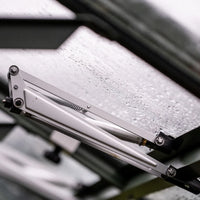 A hydraulic arm supports a rain-splattered glass pane, partially open, amidst the metal framework of a Rhino Greenhouse.