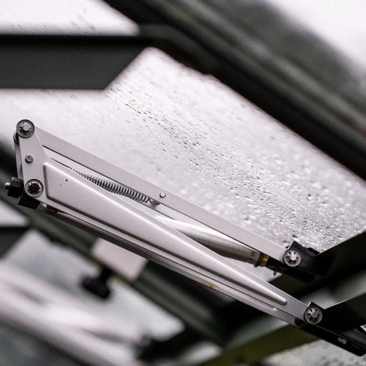 A hydraulic arm supports a rain-splattered glass pane, partially open, amidst the metal framework of a Rhino Greenhouse.