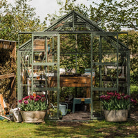 A green Rhino Greenhouse stands surrounded by lush trees, housing wooden shelves with gardening tools and pots. Two large containers of blooming tulips are positioned at the entrance.