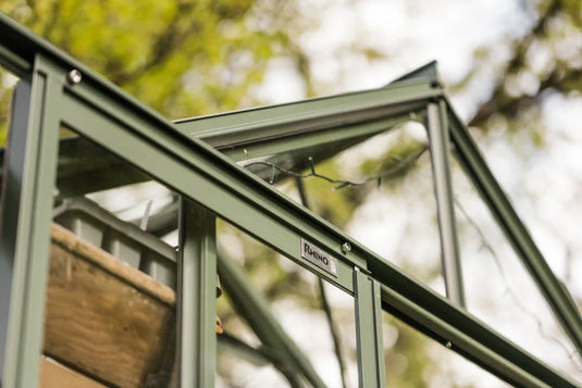 Rhino Greenhouse frame with a Rhino label, highlighting a green metal structure against a blurred background of trees and sky. A string of lights is draped along the frame.
