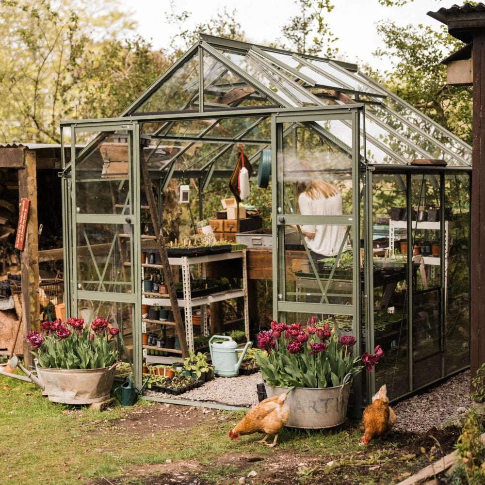 Rhino Greenhouse stands with plants and tools inside, as a person tends to seedlings. Tulips and chickens populate the grassy area outside, amidst surrounding trees and rustic structures.