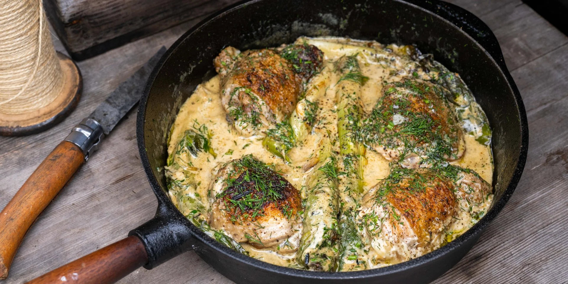A black skillet holds flavorful chicken breasts in creamy sauce, garnished with herbs. It rests on a rustic wooden surface, accompanied by a knife with a wooden handle and a spool of twine.
