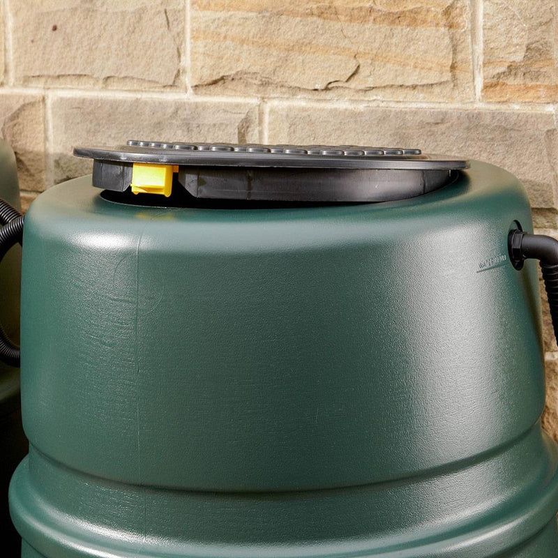 A green water butt with a black lid and yellow handle stands against a stone wall, featuring a hose connector on the side.