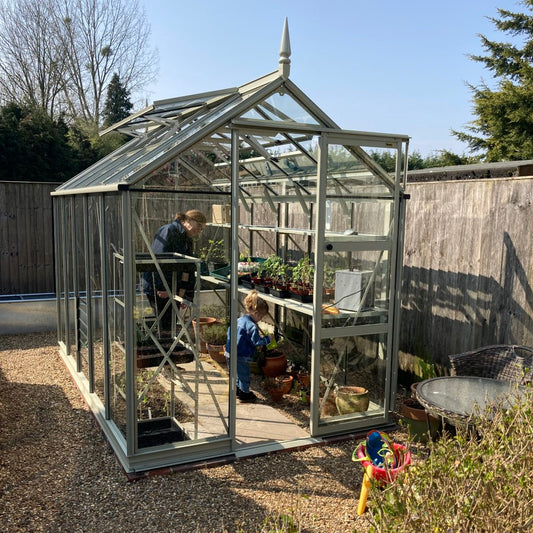 A Rhino Greenhouse contains two people tending to plants on shelves. It is situated on gravel near a wooden fence, surrounded by outdoor furniture and gardening tools.