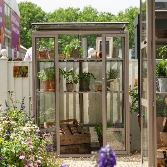 A compact Rhino Greenhouse houses potted plants, visible through glass shelves, amidst a garden with purple flowers. The Rhino Greenhouse is labeled Rhino and sits on a gravel path.