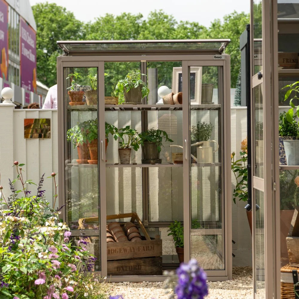 Rhino Greenhouse with potted plants inside, placed on shelves. It's situated in a garden with colorful flowers. The background features a fence and a large building sign. Text: Rhino, RIDGEWAY FARM OXFORD.