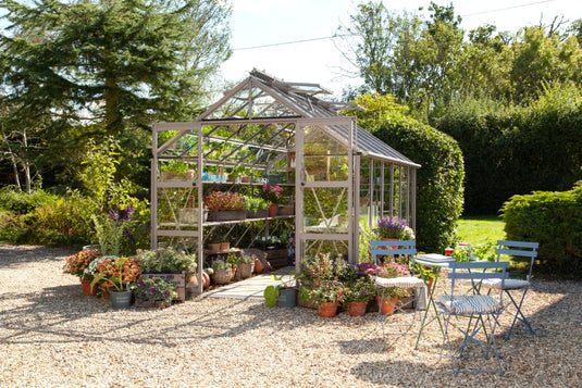 A glass Rhino Greenhouse houses assorted plants and flowers in pots, surrounded by a gravel path. Nearby are blue metallic chairs and tables set in a lush, green garden.