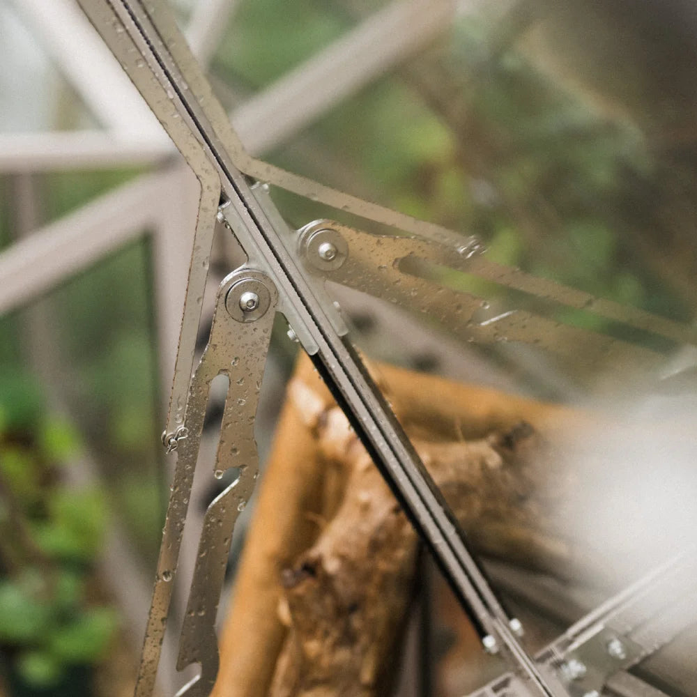 Raindrops sit on the metal framework of a Rhino Greenhouse window, secured with hinges and screws. Greenery can be seen blurred in the background, indicating a garden setting.
