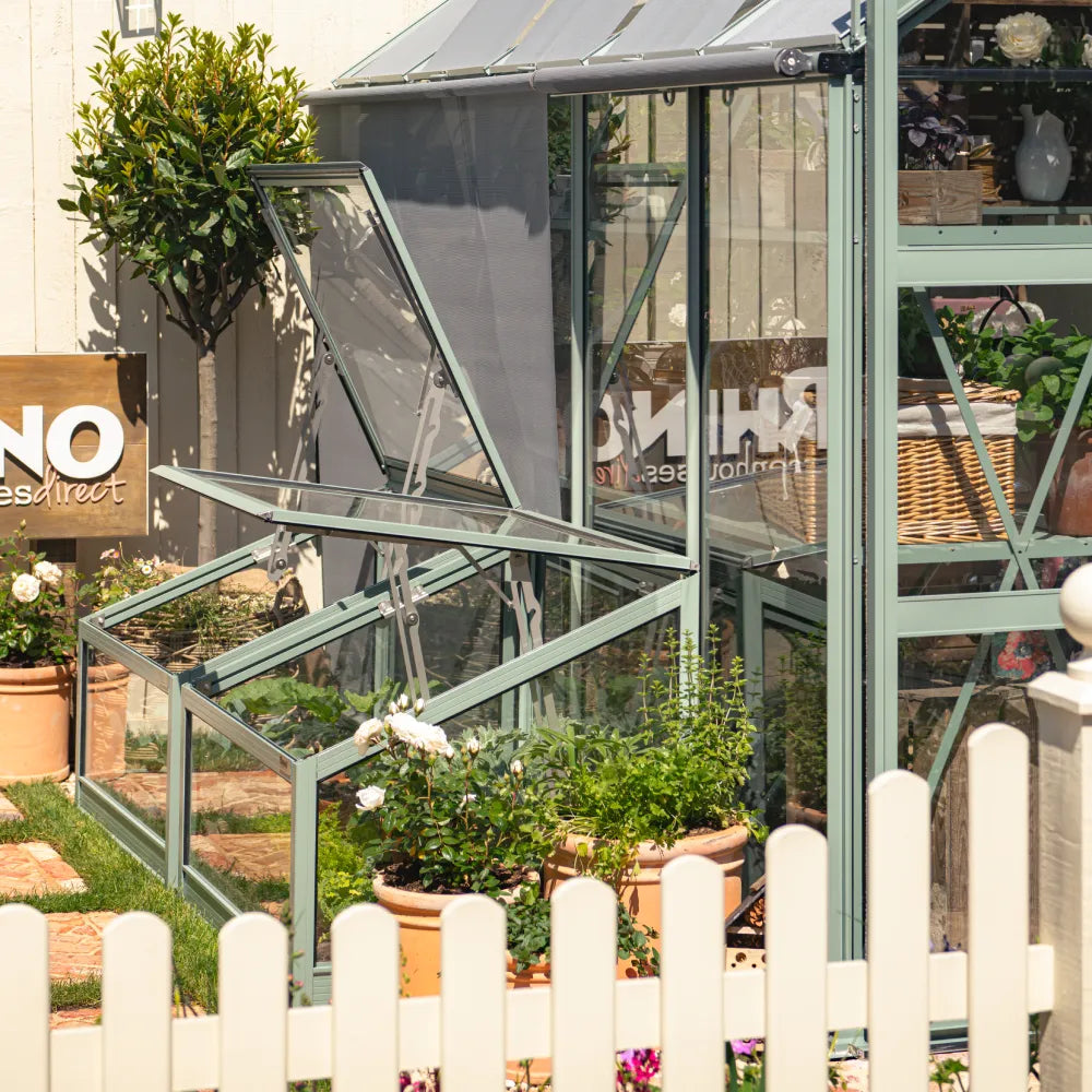 A Rhino Greenhouse with open windows allows airflow, surrounded by potted plants and flowers, within a fenced garden. A sign reads RHINO Rhino Greenhousesdirect in the background.