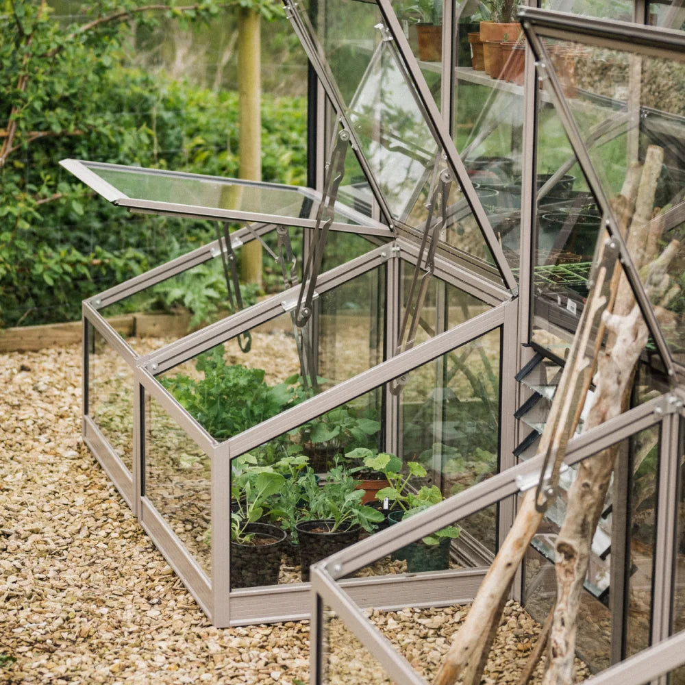 Glass Rhino Greenhouse panels are propped open, allowing ventilation around potted plants inside. The surrounding area features gravel and lush greenery, suggesting a garden setting.