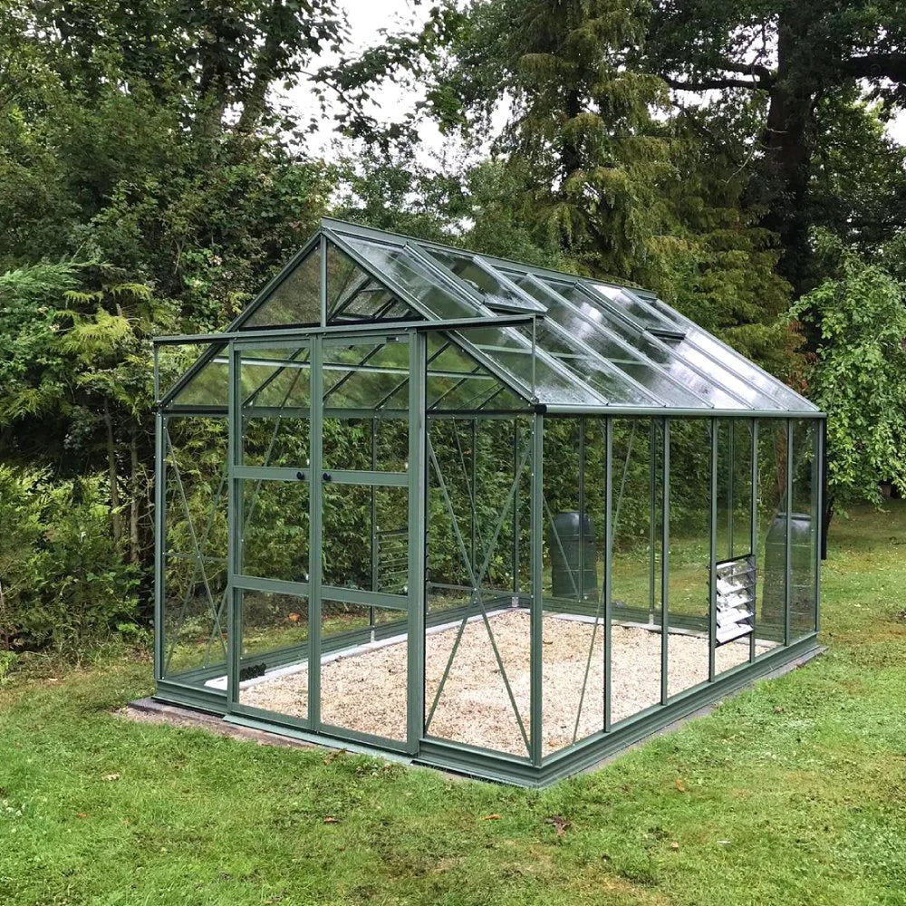 A glass Rhino Greenhouse with a green metal frame sits on a grassy lawn, surrounded by lush trees and foliage, ready for gardening use.