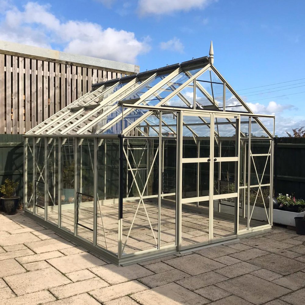 Rhino Greenhouse structure stands on a paved patio, featuring glass panels and a pointed roof accent. Wooden fencing encloses the area under a blue sky with scattered clouds.
