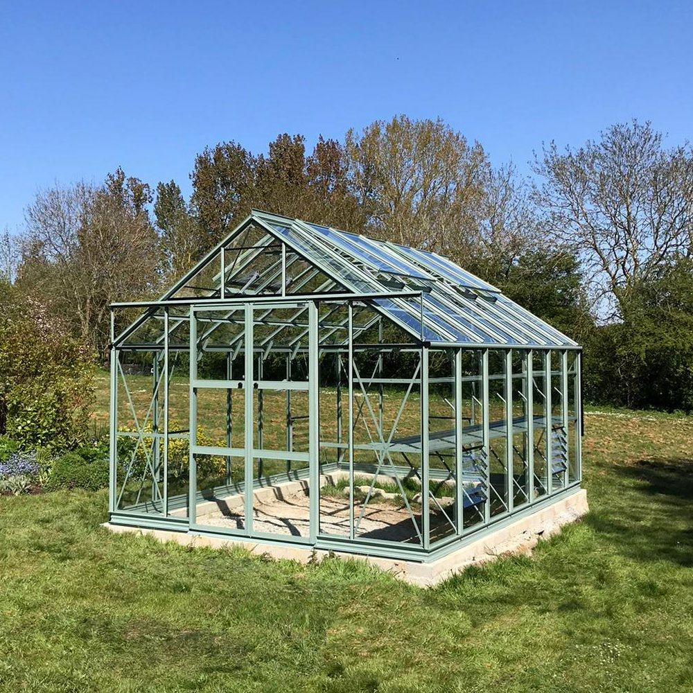 A clear, glass-paneled Rhino Greenhouse stands on grass, featuring open roof vents, surrounded by a lush garden with trees in the background under a clear blue sky.