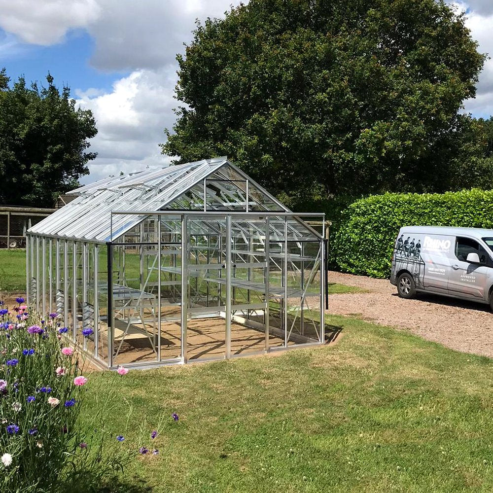 A glass Rhino Greenhouse with metal frames stands on grass, surrounded by colorful flowers. Nearby, a van with Rhino Rhino Greenhouses branding is parked, with lush trees and hedges in the background.