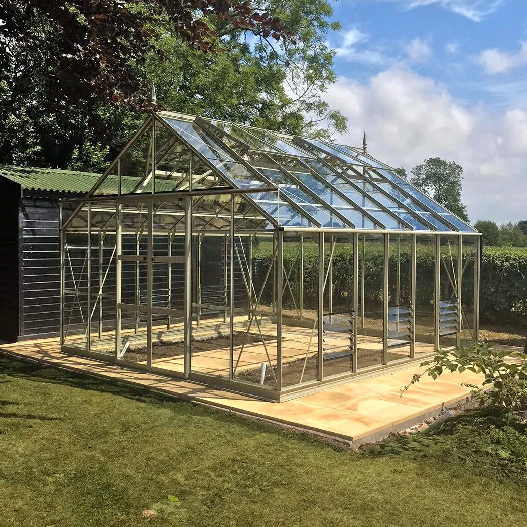 A glass and metal Rhino Greenhouse stands on a tiled base, surrounded by grass and trees, with a blue sky above.