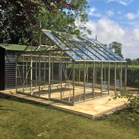 A glass and metal Rhino Greenhouse stands on a tiled base, surrounded by grass and trees, with a blue sky above.