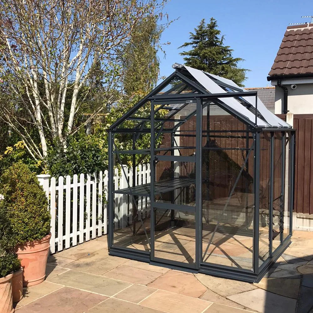 A glass and metal Rhino Greenhouse stands on a stone patio with gardening shelves inside, surrounded by a white picket fence, potted plants, and trees under a clear blue sky.