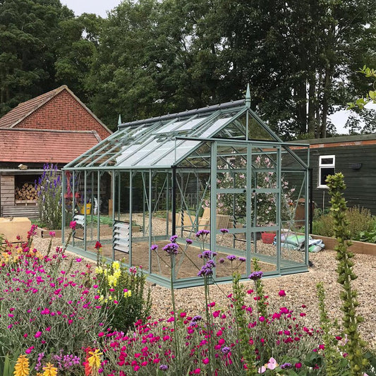 Rhino Greenhouse stands on gravel, surrounded by colorful flowering plants, with a wooden shed and trees in the background. Fiberglass panels reflect soft light, creating a serene gardening space.