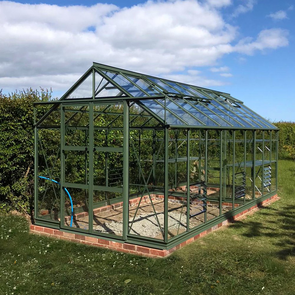 A green metal-framed Rhino Greenhouse with transparent glass panels stands in a grassy area, surrounded by hedges and under a partly cloudy sky.