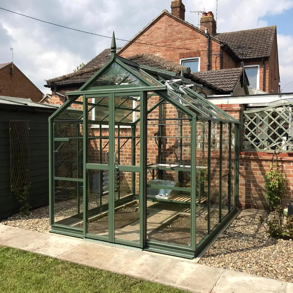 A green glass Rhino Greenhouse stands on a gravel and paved area, housing plant shelves, adjacent to a red-brick house with a pitched roof. Sky and surrounding garden visible.