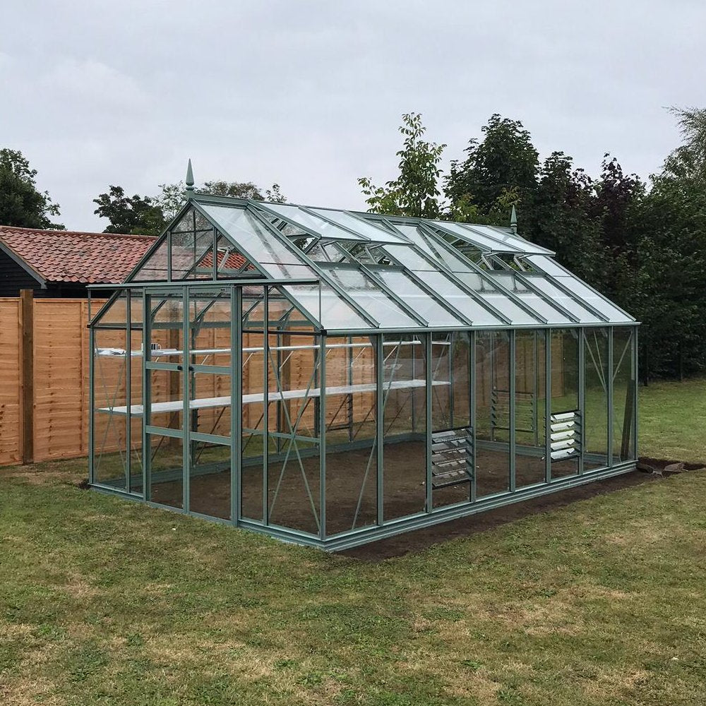 A glass and metal-framed Rhino Greenhouse stands in a grassy garden, surrounded by wooden fencing and trees, featuring empty interior shelves for plant storage and ventilation windows on the roof.