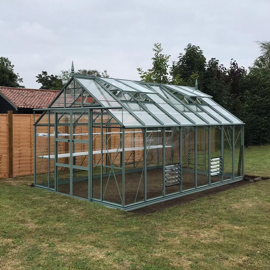 A glass and metal-framed Rhino Greenhouse stands in a grassy garden, surrounded by wooden fencing and trees, featuring empty interior shelves for plant storage and ventilation windows on the roof.