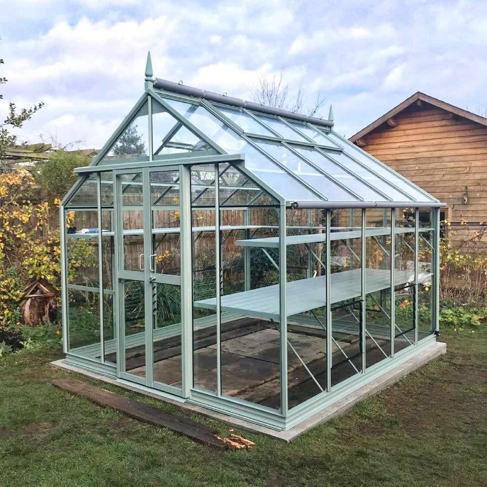 A glass Rhino Greenhouse stands in a garden, featuring a sturdy green frame and shelving. It is surrounded by grass, with a wooden shed in the background and some plants nearby.