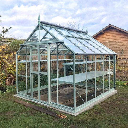A glass Rhino Greenhouse stands in a garden, featuring a sturdy green frame and shelving. It is surrounded by grass, with a wooden shed in the background and some plants nearby.