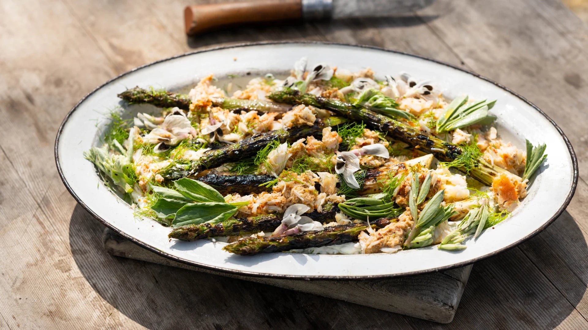 Grilled asparagus topped with herbs and shredded crab meat, arranged on a white oval plate. The dish is presented on a wooden table, with a knife in the background.