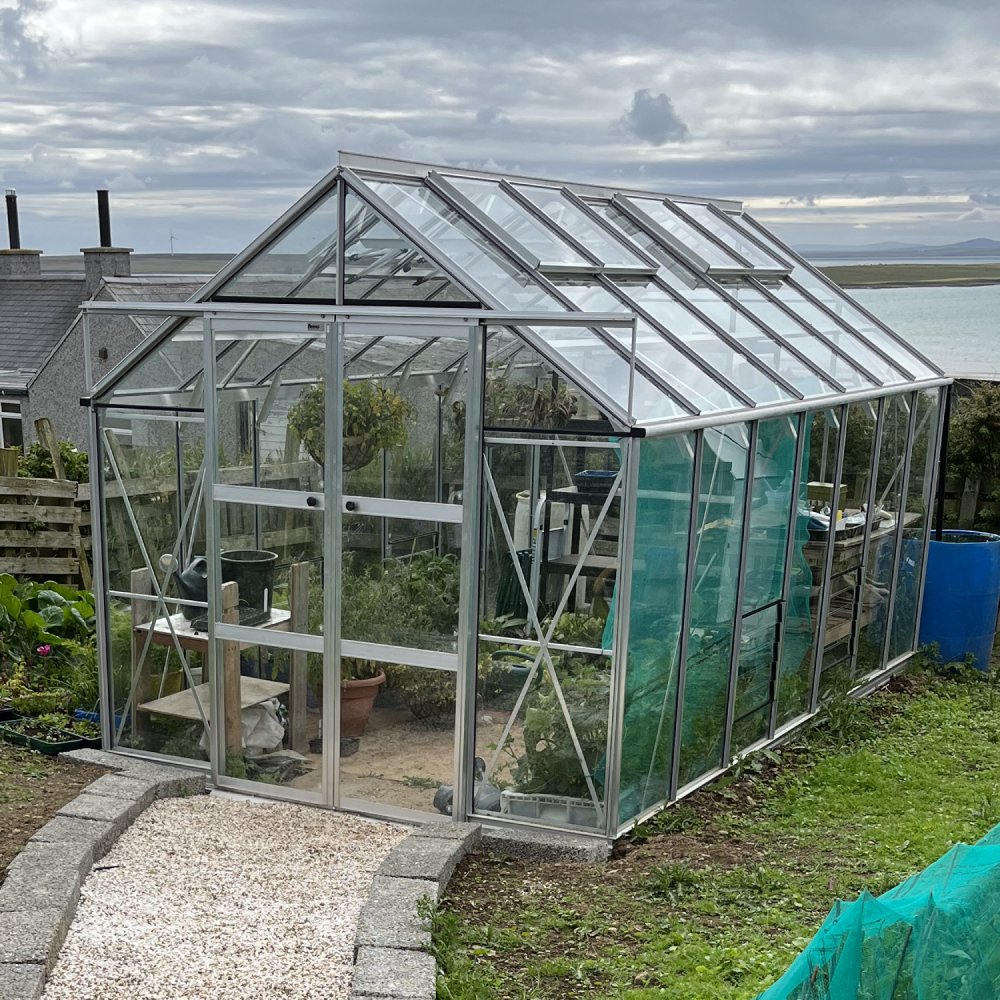 A glass Rhino Greenhouse stands on a grassy hill overlooking water. Inside, plants and gardening tools are visible. A gravel path leads to the entrance, and nearby are some outdoor structures.